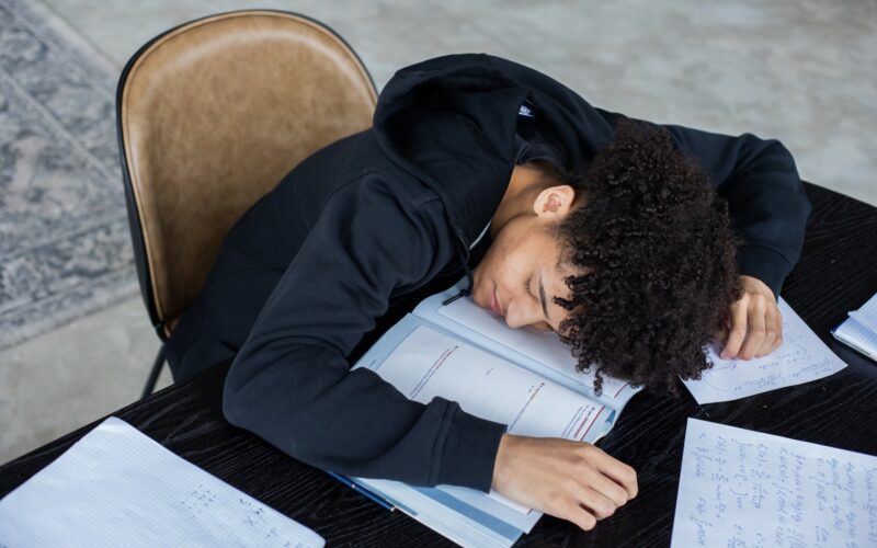 High angle of exhausted African American student resting on opened textbook and papers while preparing for exam